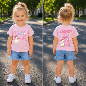 May include: A light pink short-sleeved t-shirt featuring a graphic of a duck with a pink bow. The front displays the text "TWO silly" with a heart. The back features the name "LAINEY" above a large number "2" and a matching duck graphic.