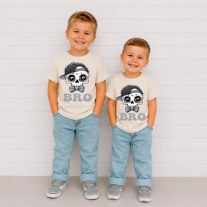 May include: Two cream-colored short-sleeve t-shirts featuring a graphic of a skull wearing a black baseball cap and a bow tie, with the word BRO printed in grey underneath. The shirts are paired with light blue denim jeans and grey sneakers.