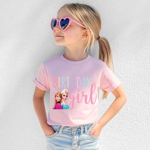K&ouml;nnte beinhalten: Ein hellrosa T-Shirt mit den Worten "BIRTHDAY girl" in einer bunten Schrift. Das Shirt zeigt eine Grafik von zwei animierten Figuren. Die Person tr&auml;gt rosa herzf&ouml;rmige Sonnenbrillen und blaue Jeans.