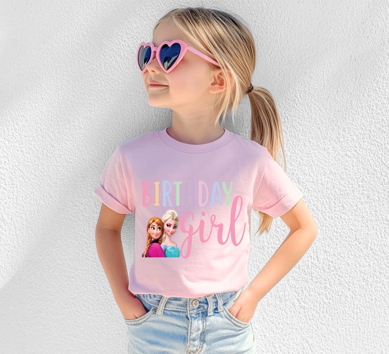 K&ouml;nnte beinhalten: Ein hellrosa T-Shirt mit den Worten "BIRTHDAY girl" in einer bunten Schrift. Das Shirt zeigt eine Grafik von zwei animierten Figuren. Die Person tr&auml;gt rosa herzf&ouml;rmige Sonnenbrillen und blaue Jeans.