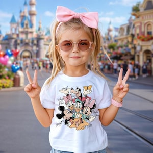 May include: A young child wearing a white t-shirt featuring a colorful graphic of Disney characters, including Mickey Mouse, Minnie Mouse, and Donald Duck. The child is also wearing pink sunglasses, a pink headband, and a pink bracelet. The child is giving the peace sign.