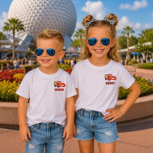 Op de afbeelding: Twee witte t-shirts met een kleine afbeelding van een rode raceauto en de tekst BRO op het ene shirt en SISSY op het andere. De shirts worden gedragen met een blauwe spijkerbroek en short, gecombineerd met pilotenbrillen en geruite haarstrikken.