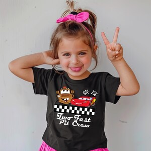 Puede incluir: Una camiseta negra con el texto "Two Fast Pit Crew" e im&aacute;genes de dibujos animados de Mater y Rayo McQueen de la pel&iacute;cula Cars. La camiseta tiene un dise&ntilde;o de bandera a cuadros y la lleva un ni&ntilde;o peque&ntilde;o.