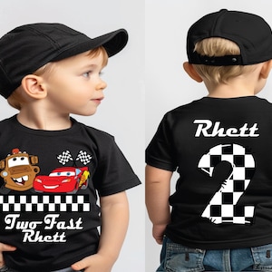 May include: Black t-shirt featuring cartoon characters Mater and Lightning McQueen with checkered flags. The shirt has the text "Two Fast Rhett" on the front and the name "Rhett" and a checkered number 2 on the back. The child is wearing a black baseball cap.