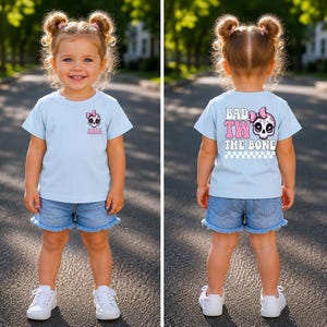 May include: A light blue t-shirt featuring a small skull graphic with a pink bow and the name Aria on the front, and a large skull graphic with a pink bow, the text BAD TO THE BONE, and a checkered pattern on the back, paired with denim shorts.