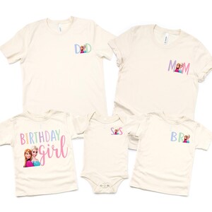 K&ouml;nnte beinhalten: Eine Sammlung cremefarbener T-Shirts und ein Body. Die Shirts haben kurze &Auml;rmel und zeigen die W&ouml;rter "DAD", "MOM", "BR" und "BIRTHDAY girl" in Pastellfarben, zusammen mit Bildern von Zeichentrickfiguren. Der Body hat die Buchstaben "S S".
