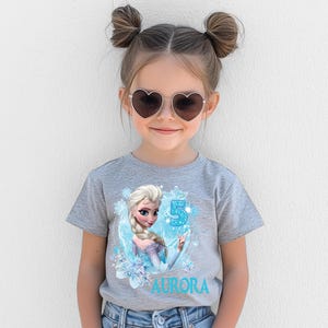 Pu&ograve; includere: Una maglietta grigia con una grafica di Elsa di Frozen, il numero 5 e il nome "Aurora" in blu. La maglietta &egrave; indossata da una bambina che indossa occhiali da sole a forma di cuore.