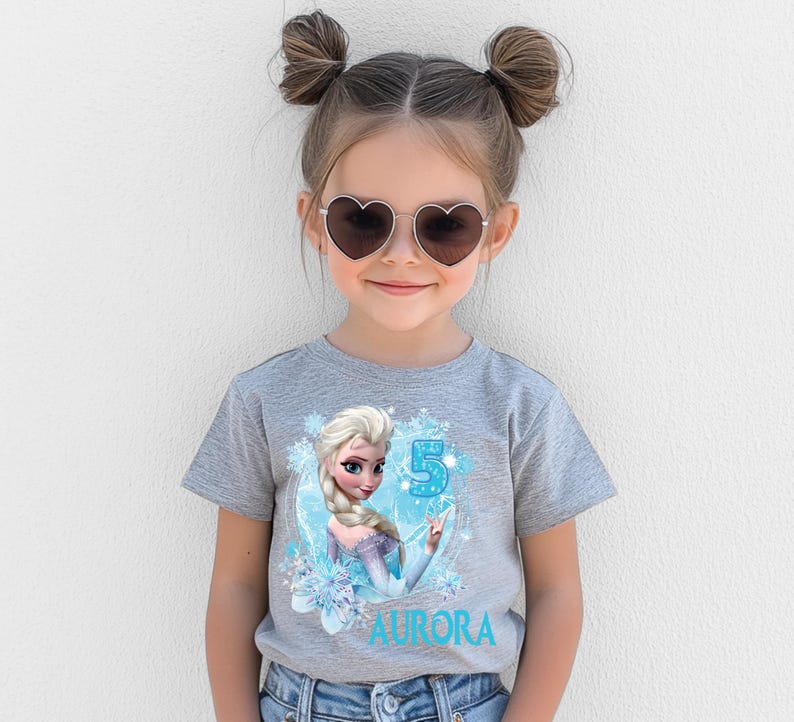 Pu&ograve; includere: Una maglietta grigia con una grafica di Elsa di Frozen, il numero 5 e il nome "Aurora" in blu. La maglietta &egrave; indossata da una bambina che indossa occhiali da sole a forma di cuore.