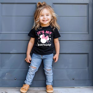 May include: A black t-shirt with the text "IT'S MY BIRTHDAY" and a white Minnie Mouse head graphic with a pink bow. The shirt is worn by a young child with blonde hair and blue jeans.