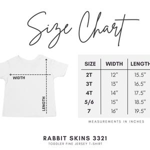 May include: A white toddler t-shirt size chart for Rabbit Skins 3321. The graphic shows a t-shirt outline with width and length arrows, and a table listing sizes 2T through 7 with corresponding measurements in inches. Text reads Size Chart.