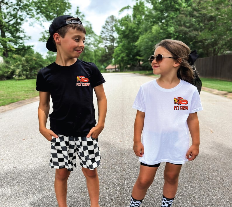 Op de afbeelding: Een zwart t-shirt en een wit t-shirt, beide met een kleine afbeelding van een rode raceauto en de tekst PIT CREW op de borst. Het zwarte shirt wordt gecombineerd met zwart-wit geblokte shorts, terwijl het witte shirt wordt gedragen met een zwarte short.