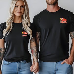Op de afbeelding: Twee zwarte T-shirts met korte mouwen en een kleine afbeelding op de linkerborst. Elk shirt toont een rode cartoon raceauto boven de tekst MAMA op het ene shirt en DADA op het andere. De shirts worden gecombineerd met een lichtblauwe spijkerbroek.