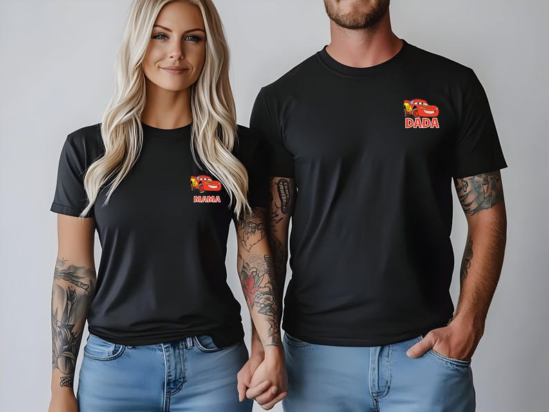Op de afbeelding: Twee zwarte T-shirts met korte mouwen en een kleine afbeelding op de linkerborst. Elk shirt toont een rode cartoon raceauto boven de tekst MAMA op het ene shirt en DADA op het andere. De shirts worden gecombineerd met een lichtblauwe spijkerbroek.