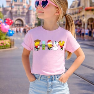May include: A young person wearing a pink t-shirt with the word "SHIBBY" in a cartoon font. The shirt is paired with denim shorts and heart-shaped sunglasses. The background features a theme park.