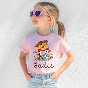 K&ouml;nnte beinhalten: Hellrosa T-Shirt mit einer bunten Grafik von Toy Story-Charakteren. Die Zahl "2" ist mit einem Cowboyhut und Halstuch gestaltet. Der Name "Sadie" steht in Schreibschrift unter der Grafik.