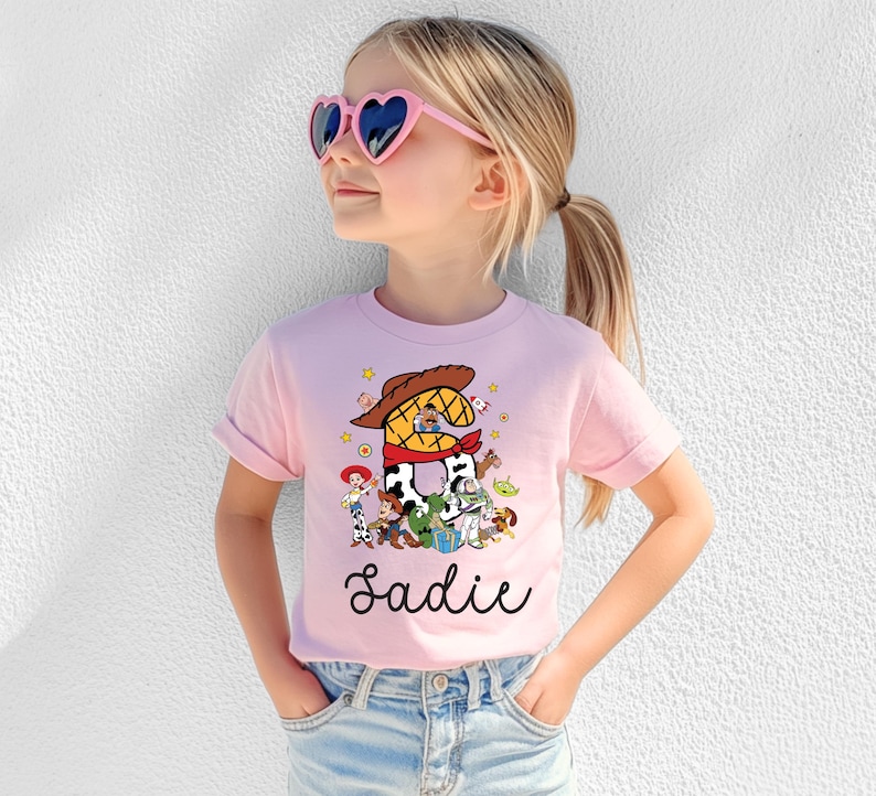 K&ouml;nnte beinhalten: Hellrosa T-Shirt mit einer bunten Grafik von Toy Story-Charakteren. Die Zahl "2" ist mit einem Cowboyhut und Halstuch gestaltet. Der Name "Sadie" steht in Schreibschrift unter der Grafik.