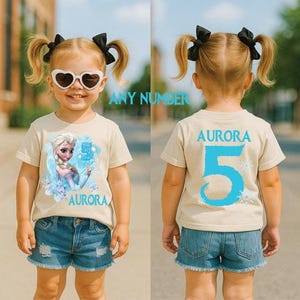Pu&ograve; includere: Una maglietta beige con una grafica di Elsa di Frozen, il numero 5 e il nome "Aurora" in blu. Il retro della maglietta mostra anche "Aurora" e un grande 5 blu. La bambina indossa pantaloncini di jeans.