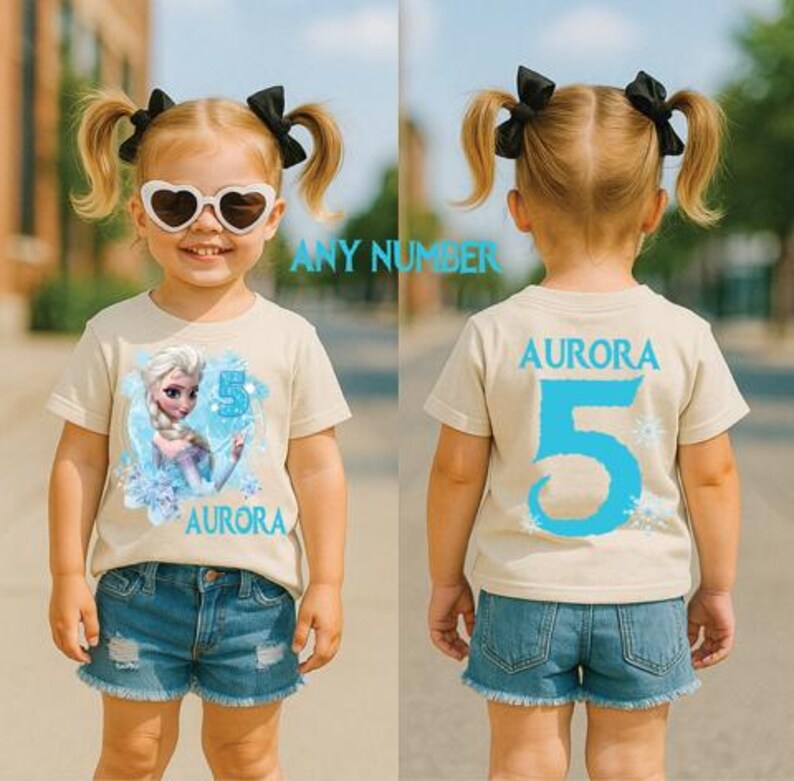 Pu&ograve; includere: Una maglietta beige con una grafica di Elsa di Frozen, il numero 5 e il nome "Aurora" in blu. Il retro della maglietta mostra anche "Aurora" e un grande 5 blu. La bambina indossa pantaloncini di jeans.