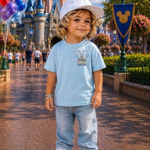 Pu&ograve; includere: Un bambino indossa una maglietta azzurra con un disegno del castello Disney e il nome "DEAN". Indossa anche jeans azzurri, scarpe da ginnastica bianche e un berretto bianco. Lo sfondo presenta un castello e palloncini.