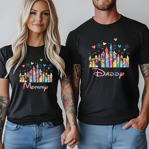 May include: Black t-shirts with a colorful castle design and the words "Mommy" and "Daddy" in a playful font. The design includes various cartoon characters and castle turrets. The shirts are worn by two people.