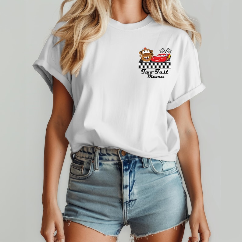 Puede incluir: Camiseta blanca con un dise&ntilde;o de dos coches de carreras de dibujos animados y el texto "Two Fast Mama" en negro. El dise&ntilde;o incluye una bandera a cuadros y un borde a cuadros blanco y negro.