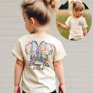 Pu&ograve; includere: Una maglietta beige con una grafica colorata di un castello, un fiocco e orecchie di Topolino, con la scritta "Disney Trip 2026". La maglietta &egrave; visibile su un bambino piccolo.