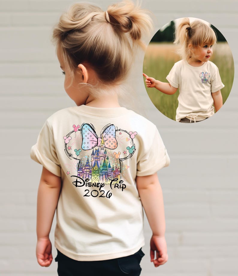 Pu&ograve; includere: Una maglietta beige con una grafica colorata di un castello, un fiocco e orecchie di Topolino, con la scritta "Disney Trip 2026". La maglietta &egrave; visibile su un bambino piccolo.