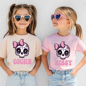 May include: A beige t-shirt with a pink skull graphic wearing a bow and the text COUSIN, and a light pink t-shirt with a matching skull graphic and the text SISSY. Both shirts feature a cartoon-style skull with large eyes and a pink bow.