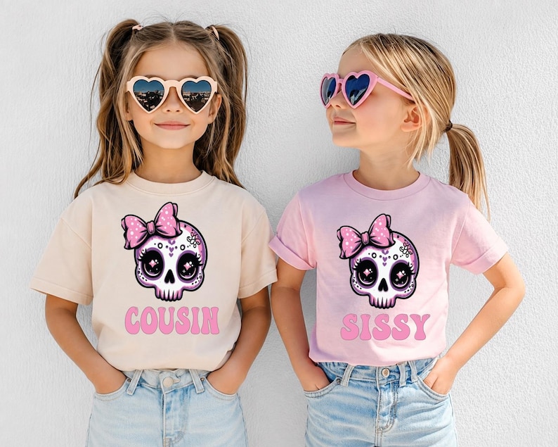 May include: A beige t-shirt with a pink skull graphic wearing a bow and the text COUSIN, and a light pink t-shirt with a matching skull graphic and the text SISSY. Both shirts feature a cartoon-style skull with large eyes and a pink bow.