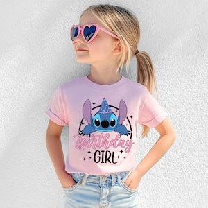 May include: A light pink t-shirt featuring a blue cartoon character wearing a party hat, with the words "Birthday Girl" in pink script. The shirt is worn by a young girl wearing heart-shaped sunglasses.