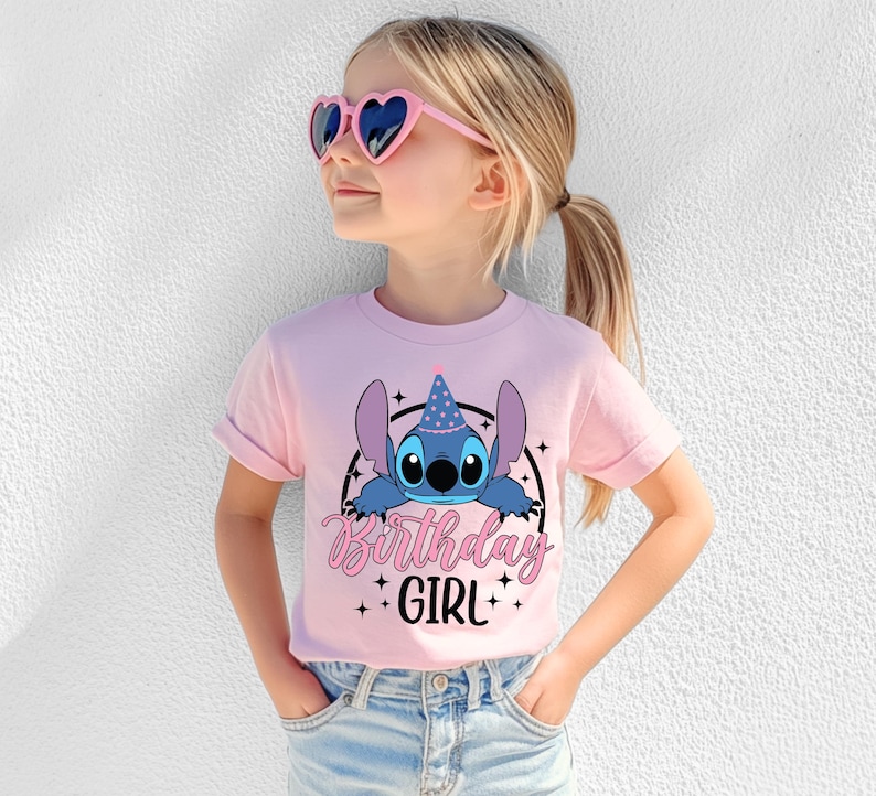 May include: A light pink t-shirt featuring a blue cartoon character wearing a party hat, with the words "Birthday Girl" in pink script. The shirt is worn by a young girl wearing heart-shaped sunglasses.