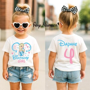 May include: A white short-sleeved t-shirt featuring a graphic of a blonde character in a blue dress with the text Birthday Girl on the front and the name Daphne with the number 4 on the back, paired with blue denim shorts and a black and white bow.