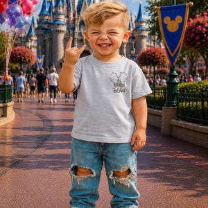 Pu&ograve; includere: Un bambino indossa una maglietta grigia con un disegno di castello colorato e il nome "DEAN" stampato sotto. Il bambino indossa jeans blu strappati e scarpe da ginnastica bianche, sorride e fa un gesto rock and roll con la mano. Palloncini e un castello sullo sfondo.