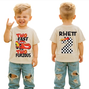 May include: A tan short-sleeved t-shirt with the text "TWO FAST TWO FURIOUS" and a red race car graphic. The back of the shirt has the name "RHETT" and a number "2" with a checkered flag design. The child is wearing ripped blue jeans.