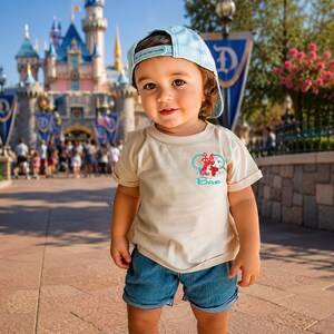 May include: A light blue baseball cap and a cream-colored short-sleeve t-shirt featuring a graphic of Ariel from The Little Mermaid with a red bow and the text Bro in blue script. The outfit includes blue denim shorts with a cuffed hem.
