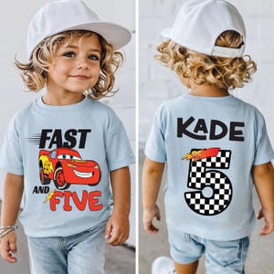 K&ouml;nnte beinhalten: Ein hellblaues T-Shirt mit einer roten Rennwagen-Grafik und dem Text FAST AND FIVE auf der Vorderseite sowie dem Namen KADE mit der Zahl 5 im Schachbrettmuster auf der R&uuml;ckseite, kombiniert mit einer wei&szlig;en Baseballkappe und hellblauer Jeans.