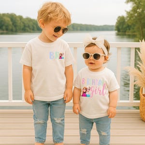K&ouml;nnte beinhalten: Zwei Kinder in wei&szlig;en T-Shirts und blauen Jeans. Die Shirts zeigen die Aufschrift "BR" und "Birthday Girl" mit einem Cartoon-Bild. Beide Kinder tragen Sonnenbrillen. Im Hintergrund sind ein See und ein wei&szlig;es Gel&auml;nder zu sehen.