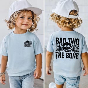 May include: A light blue short-sleeve t-shirt featuring a small black skull and checkerboard graphic with the name HENDREX on the front. The back displays a large black graphic of a skull, skeleton hands, and the text BAD TWO THE BONE.
