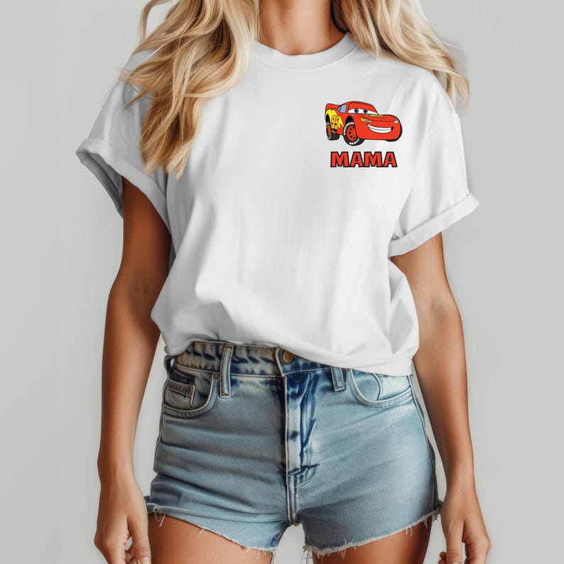 Op de afbeelding: Een wit T-shirt met korte mouwen en een kleine afbeelding op de linkerborst. De afbeelding toont een rode cartoonraceauto met een geel bliksemontwerp, geplaatst boven de rode tekst MAMA in een vetgedrukt lettertype.
