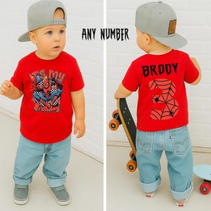 May include: A toddler in a red t-shirt featuring a Spider-Man design and the words "It's My Birthday". The child wears light blue jeans and a grey baseball cap. The back of the shirt says "Broddy" and the number "3".