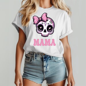 May include: A white short-sleeve t-shirt featuring a graphic of a stylized pink and white skull with large dark eyes and a pink polka-dot bow. Below the skull, the word MAMA is printed in a bold, pink, retro-style font on the front of the shirt.