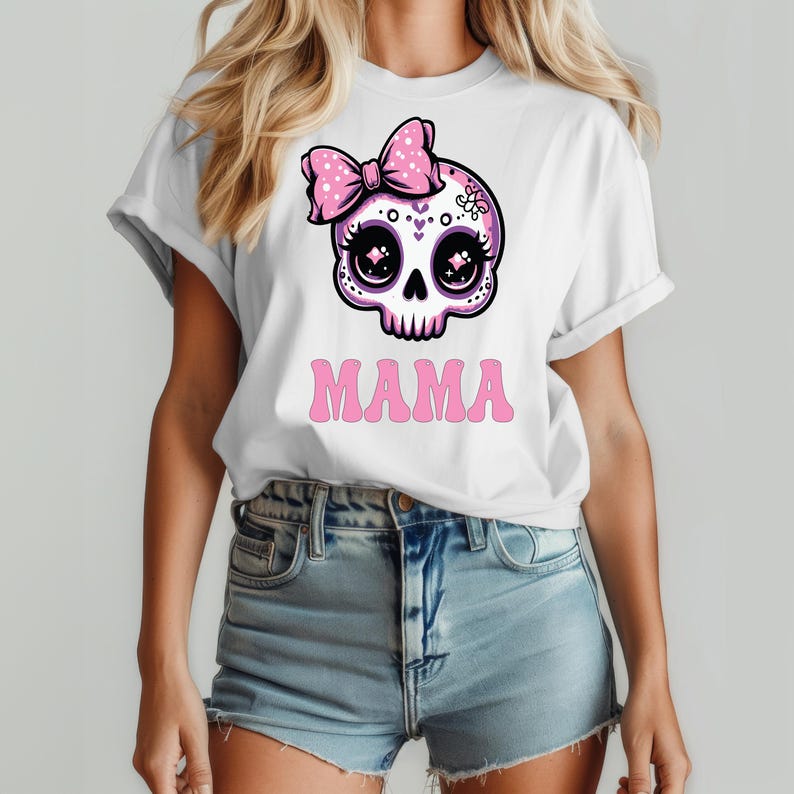 May include: A white short-sleeve t-shirt featuring a graphic of a stylized pink and white skull with large dark eyes and a pink polka-dot bow. Below the skull, the word MAMA is printed in a bold, pink, retro-style font on the front of the shirt.