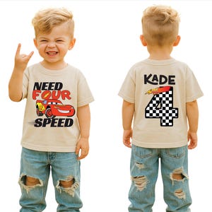 Op de afbeelding: Een beige t-shirt met korte mouwen en een afbeelding van een rode raceauto met de tekst "NEED FOUR SPEED" op de voorkant. De achterkant toont de naam "KADE" boven een groot cijfer "4" met een zwart-wit geblokt patroon en een bliksemschicht.