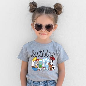 K&ouml;nnte beinhalten: Ein graues T-Shirt mit dem Schriftzug "Birthday Girl" in schwarzer Schrift und bunten Zeichentrickfiguren. Das Shirt wird von einem jungen M&auml;dchen mit herzf&ouml;rmiger Sonnenbrille getragen.