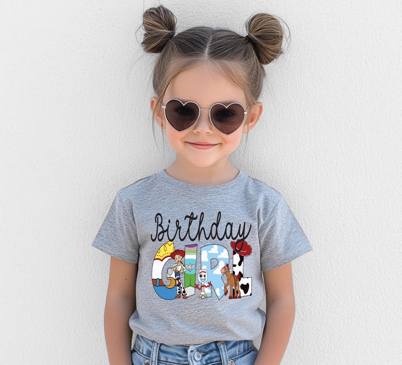 K&ouml;nnte beinhalten: Ein graues T-Shirt mit dem Schriftzug "Birthday Girl" in schwarzer Schrift und bunten Zeichentrickfiguren. Das Shirt wird von einem jungen M&auml;dchen mit herzf&ouml;rmiger Sonnenbrille getragen.