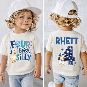 May include: A cream-colored t-shirt featuring a blue graphic of a duck wearing a cap and the text Four ever Silly on the front. The back displays the name RHETT, a large number 4, and a matching duck graphic with blue stars and accents.