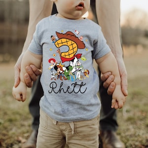 K&ouml;nnte beinhalten: Ein graues Kurzarm-T-Shirt mit einer bunten Grafik von Toy Story-Charakteren. Die Zahl 3 ist prominent dargestellt, zusammen mit dem Namen "Rhett" in Schreibschrift. Das Kind tr&auml;gt eine khakifarbene Hose.