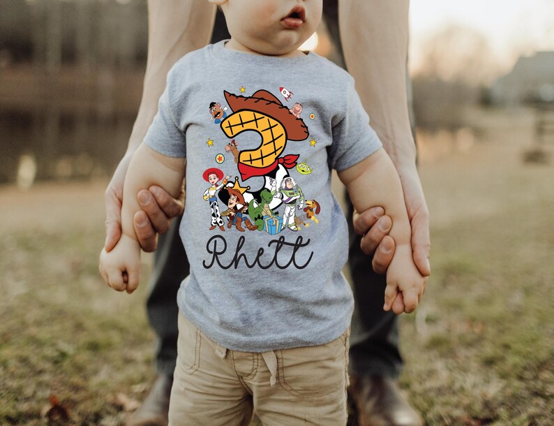 K&ouml;nnte beinhalten: Ein graues Kurzarm-T-Shirt mit einer bunten Grafik von Toy Story-Charakteren. Die Zahl 3 ist prominent dargestellt, zusammen mit dem Namen "Rhett" in Schreibschrift. Das Kind tr&auml;gt eine khakifarbene Hose.
