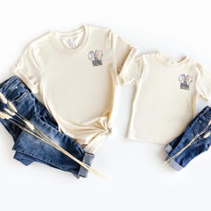 Pu&ograve; includere: Due magliette color crema, una per adulti e una per bambini, con una grafica colorata e la scritta "SWEETS". Abbinate a jeans blu e fiori secchi. Un outfit casual e coordinato.