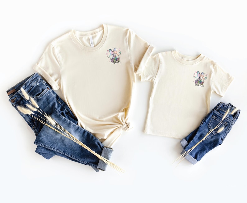 Pu&ograve; includere: Due magliette color crema, una per adulti e una per bambini, con una grafica colorata e la scritta "SWEETS". Abbinate a jeans blu e fiori secchi. Un outfit casual e coordinato.